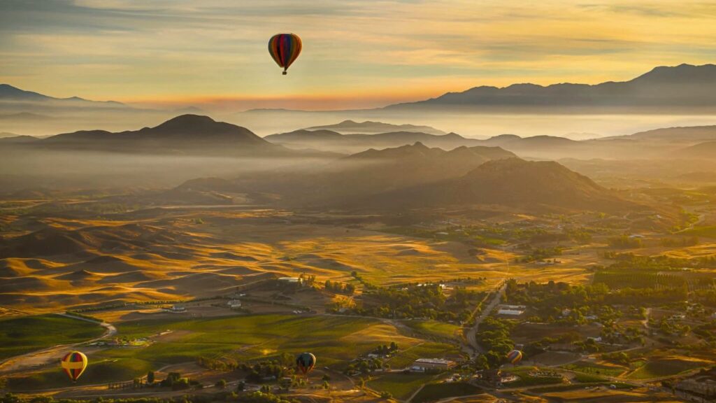 Hot air balloons floating over Temecula Valley at sunrise, with rolling hills, vineyards, and distant mountains bathed in golden morning light.